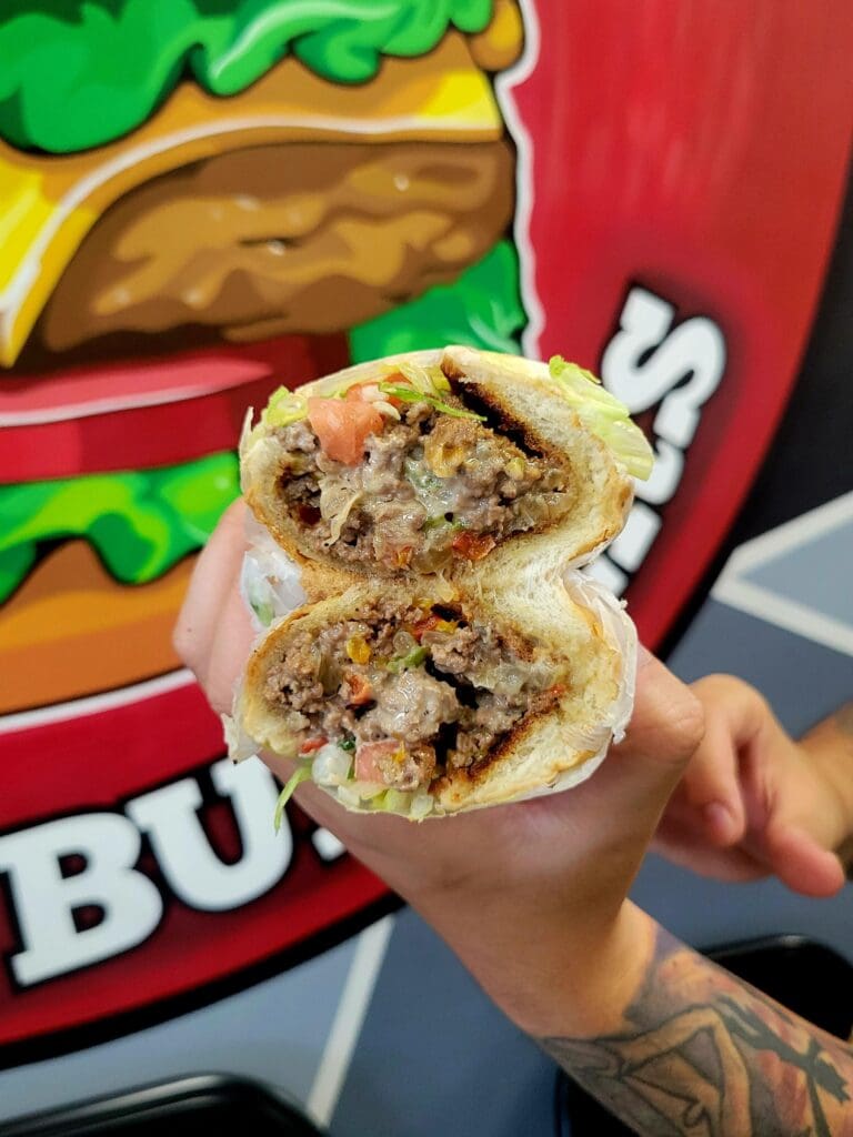 A person holds up a cross-sectioned burrito wrapped in paper against a colorful fast-food restaurant backdrop with branded signage.