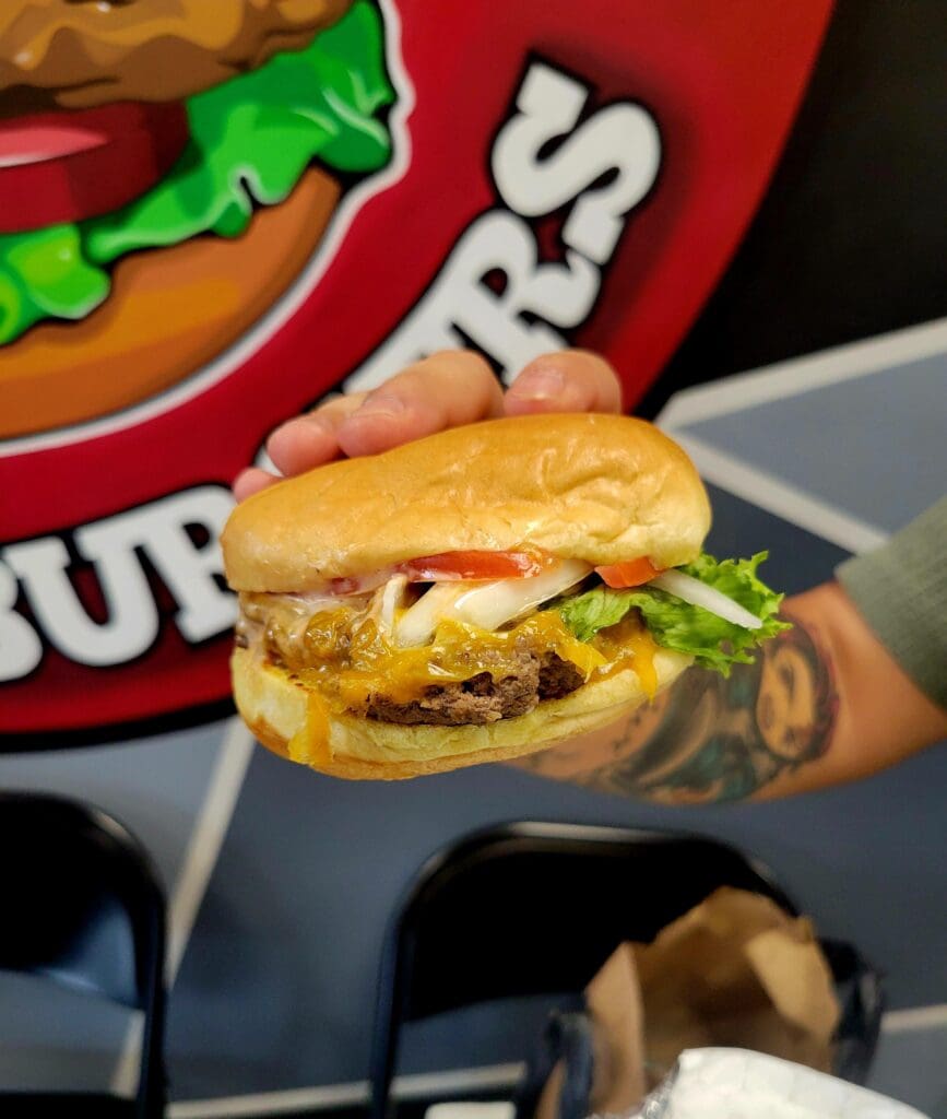 A close-up shot of a gourmet cheeseburger with melted cheese and fresh greens, displayed against branded fast-food restaurant signage.