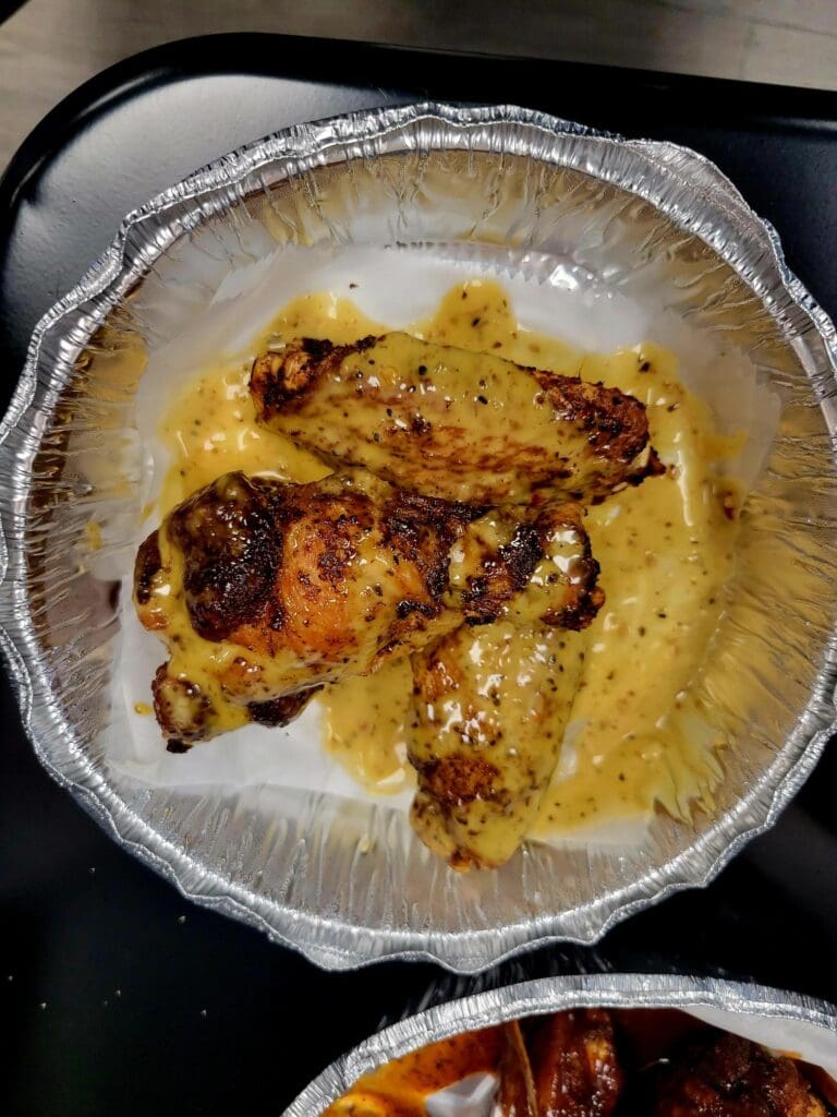 A close-up overhead shot of roasted chicken pieces in an aluminum foil takeout container with golden-brown seasoning and sauce.