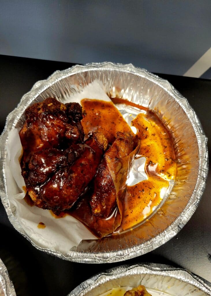A close-up overhead view of grilled ribs and chicken served in an aluminum foil container at what appears to be a food service or barbecue establishment.