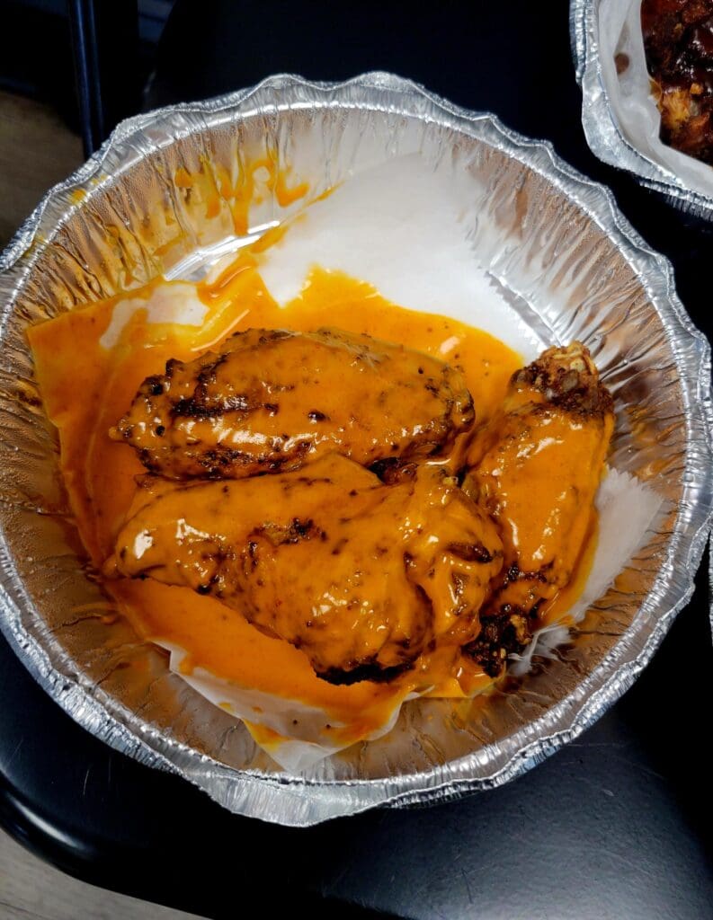 A close-up overhead view of cooked chicken pieces in an orange/golden sauce served in a disposable aluminum foil container.
