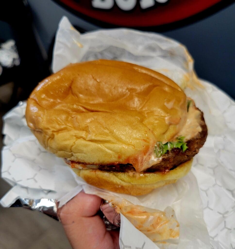 A hand holding a golden-brown breakfast sandwich wrapped in paper, featuring what appears to be bacon, lettuce, and other fillings on a toasted bun.