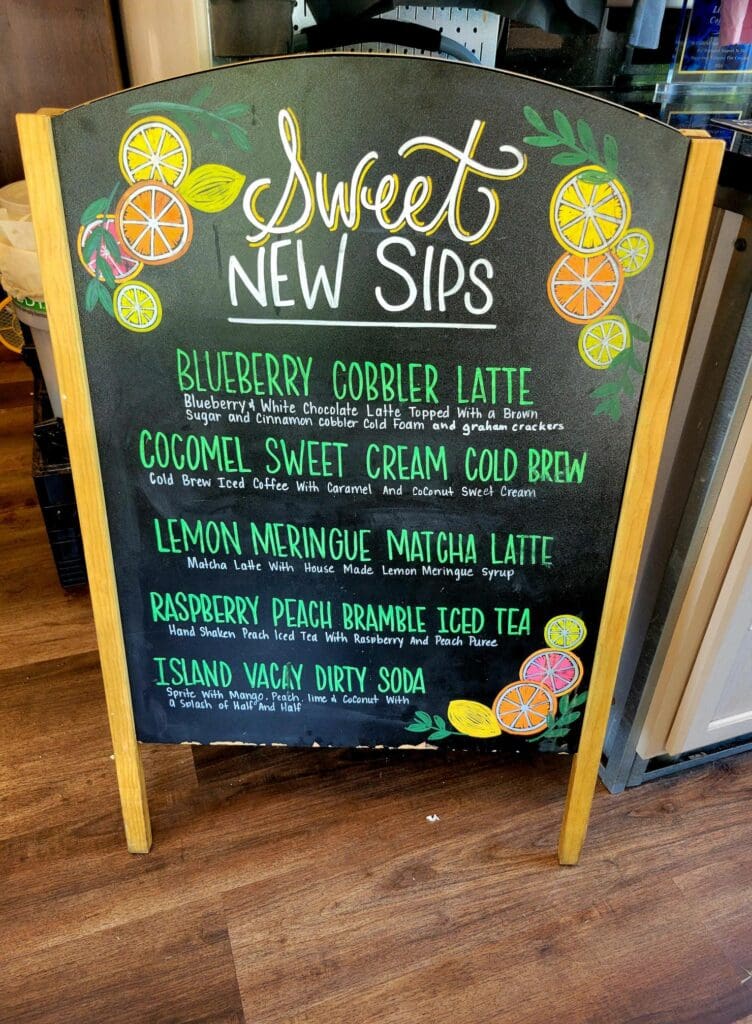 A chalkboard menu displays colorful specialty beverages with hand-drawn illustrations and decorative citrus slices against a dark green background.