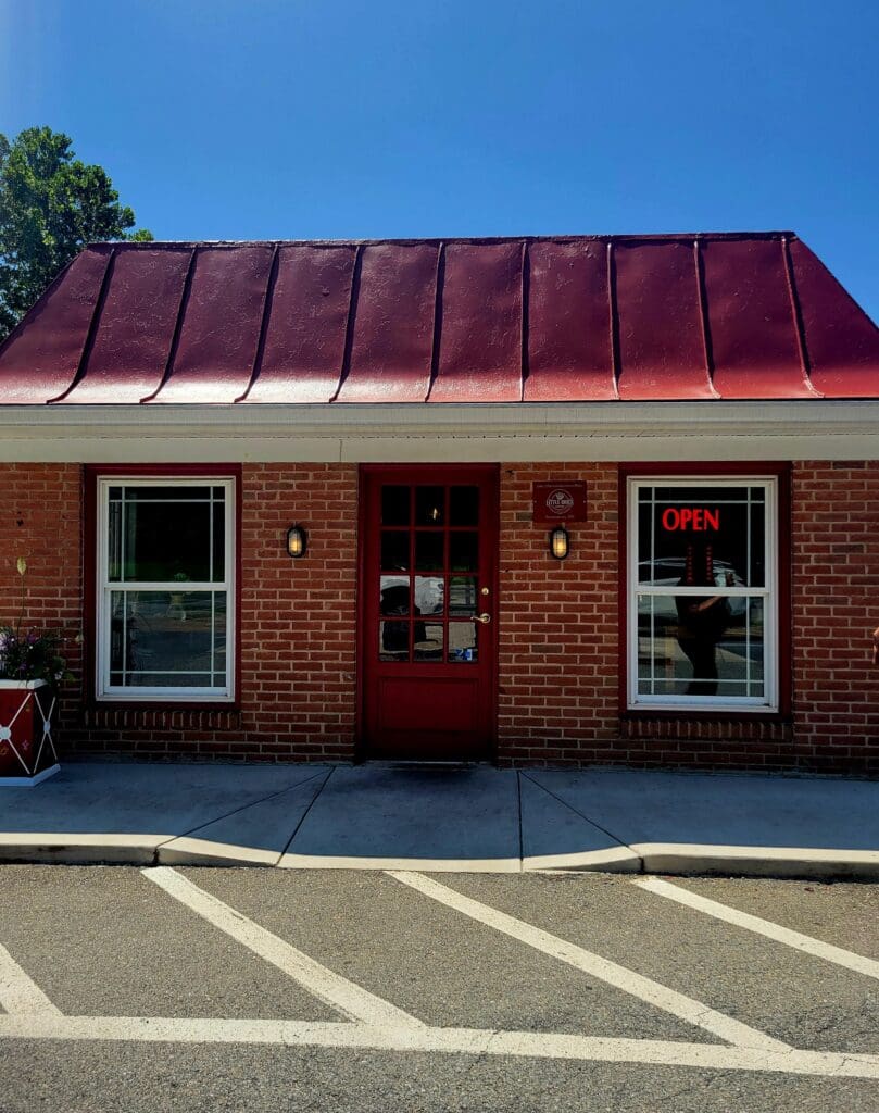 A brick storefront with a distinctive burgundy metal roof sits in a sunny parking lot with clear striped markings.