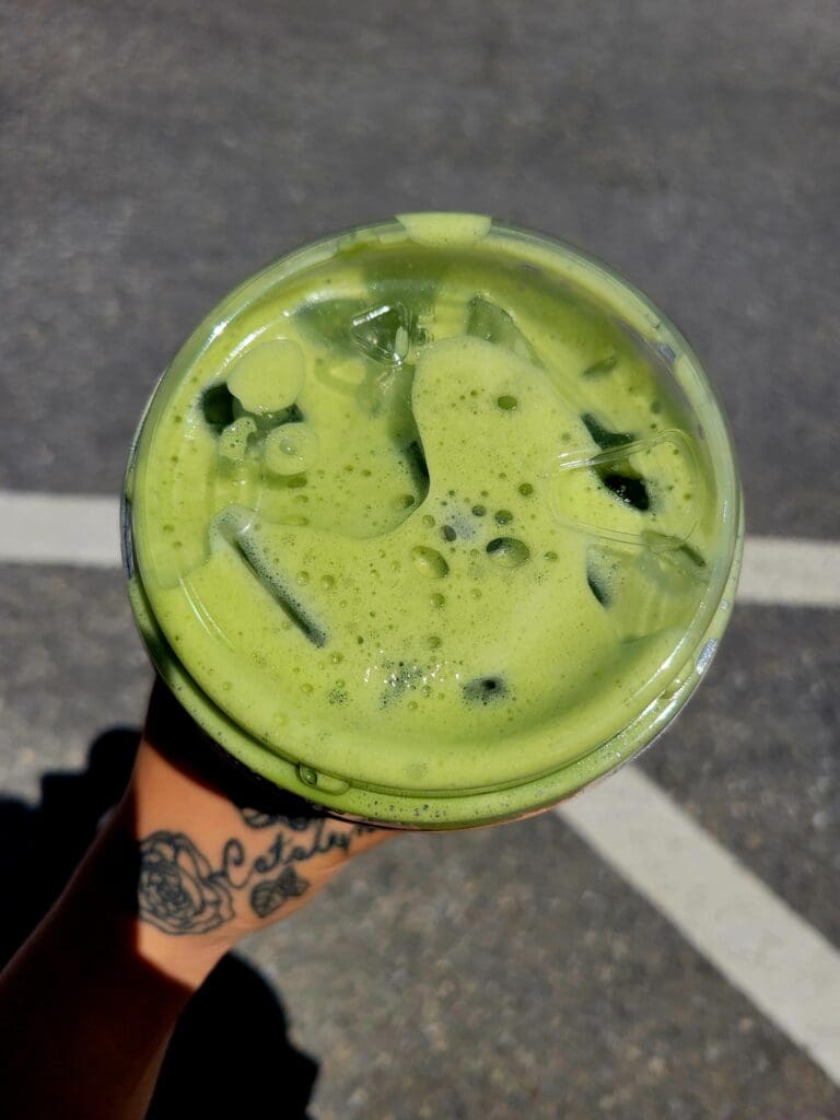 A close-up overhead shot of a vibrant green matcha or matcha-based beverage held by a person with a visible tattoo on an asphalt surface.