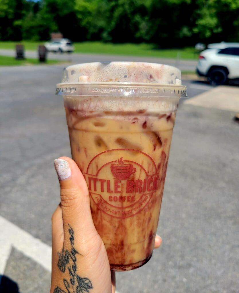 A person holds a clear cup of iced tea or cola drink with a red logo visible on it in an outdoor parking lot on a sunny day.