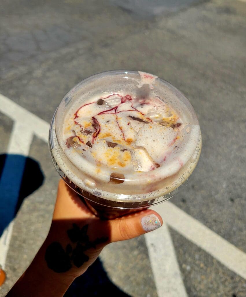 A close-up shot of a hand holding a clear plastic cup filled with a dark iced beverage with cream swirls, photographed outdoors on asphalt.