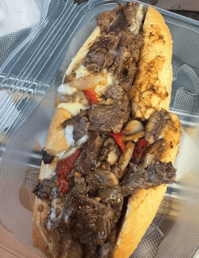 A close-up shot of a stacked philly cheesesteak sandwich with melted cheese, sliced meat, and peppers served on a clear plastic tray.