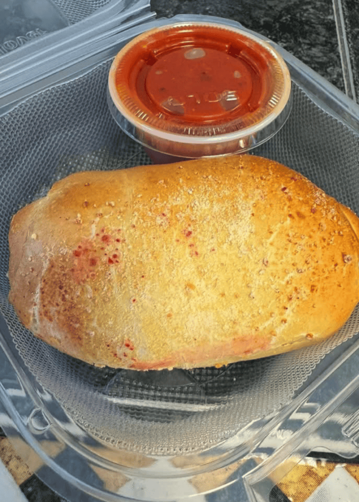 A freshly baked piece of golden cornbread sits in a clear plastic container with a small cup of red sauce on the side.