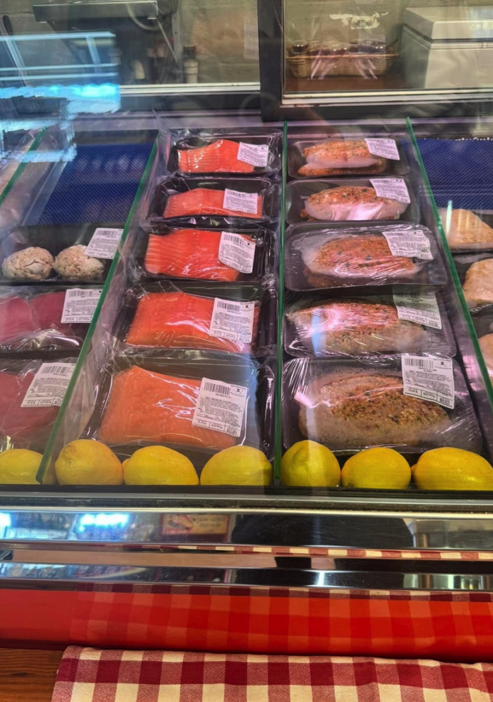 A refrigerated butcher shop display case showcasing various cuts of red meat packaged in trays, with fresh yellow lemons arranged at the bottom and a checkered cloth visible at the base.