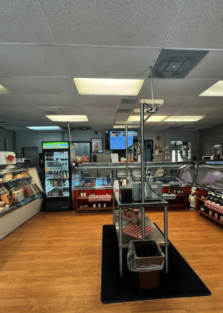 An empty convenience store or gas station interior with refrigerated displays, product shelving, fluorescent ceiling lights, and wooden flooring.