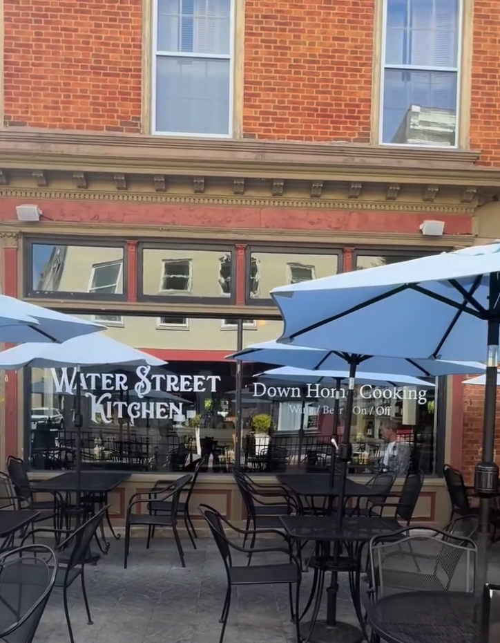 An inviting outdoor dining patio fronts a brick building with blue umbrellas shading empty tables and chairs, featuring storefront signage for Water Street Kitchen.