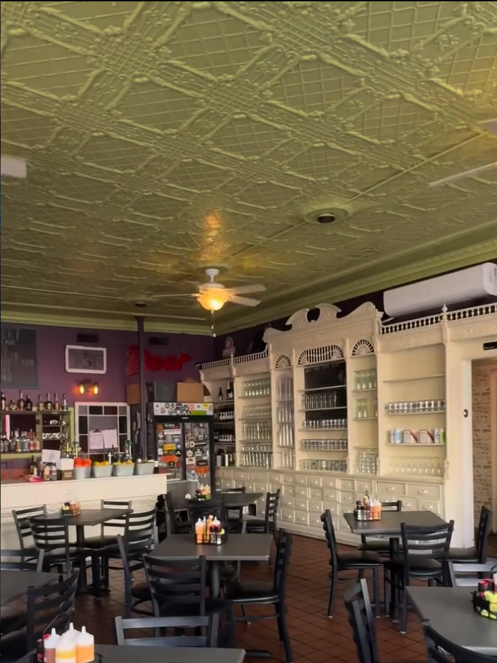 An upscale restaurant interior with sage green tin ceiling, white built-in shelving, black dining tables and chairs, and warm ambient lighting creating an elegant dining atmosphere.