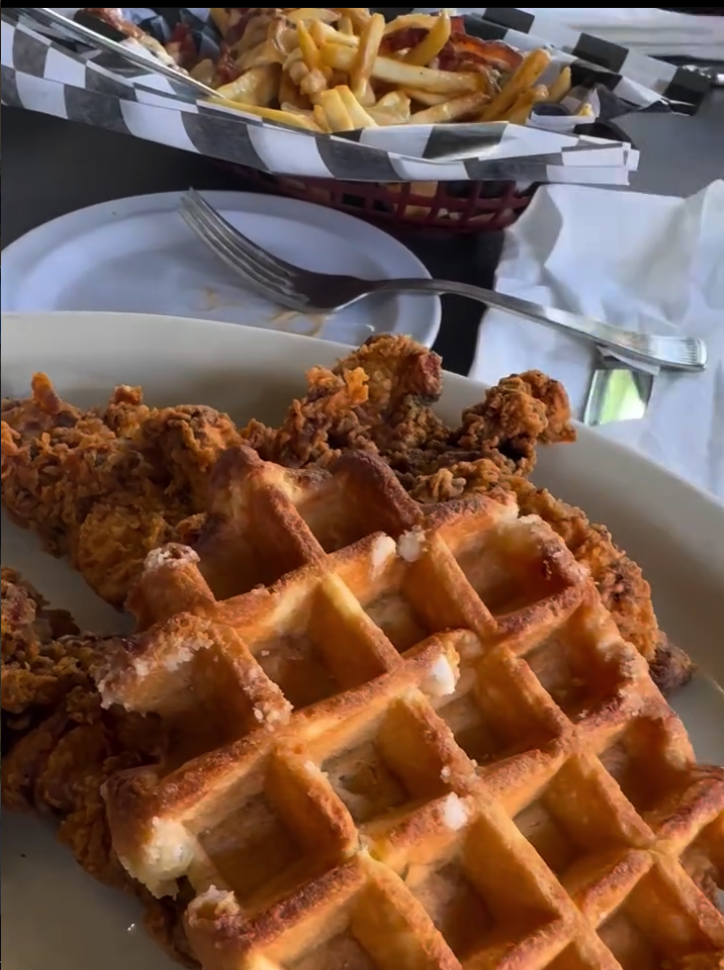 A crispy fried chicken waffle is served in a white paper container alongside golden french fries in a carnival or food service setting.