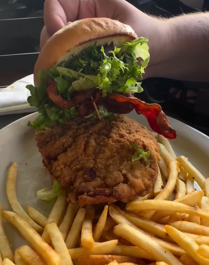 A person holds a crispy golden fried chicken burger topped with fresh lettuce and tomato, served with a side of golden french fries on a white plate.