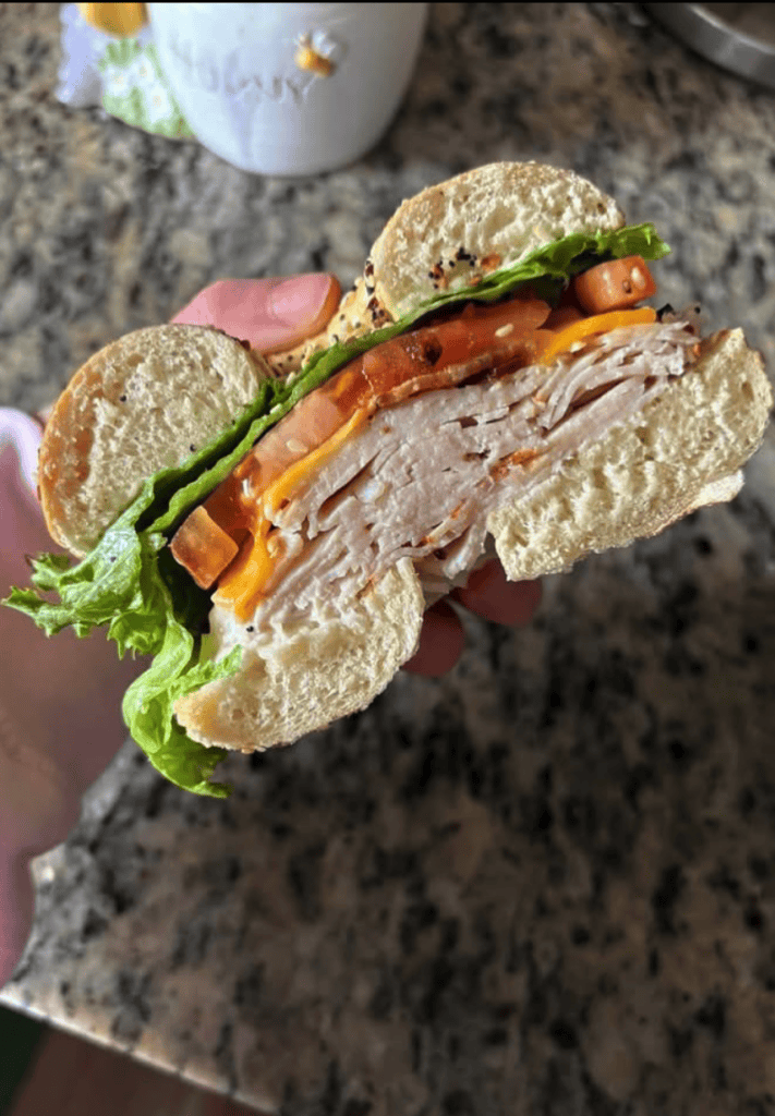 A close-up shot of a freshly made sandwich with lettuce, tomato, and other vegetables on whole wheat bread being held by someone's hand over a granite countertop.