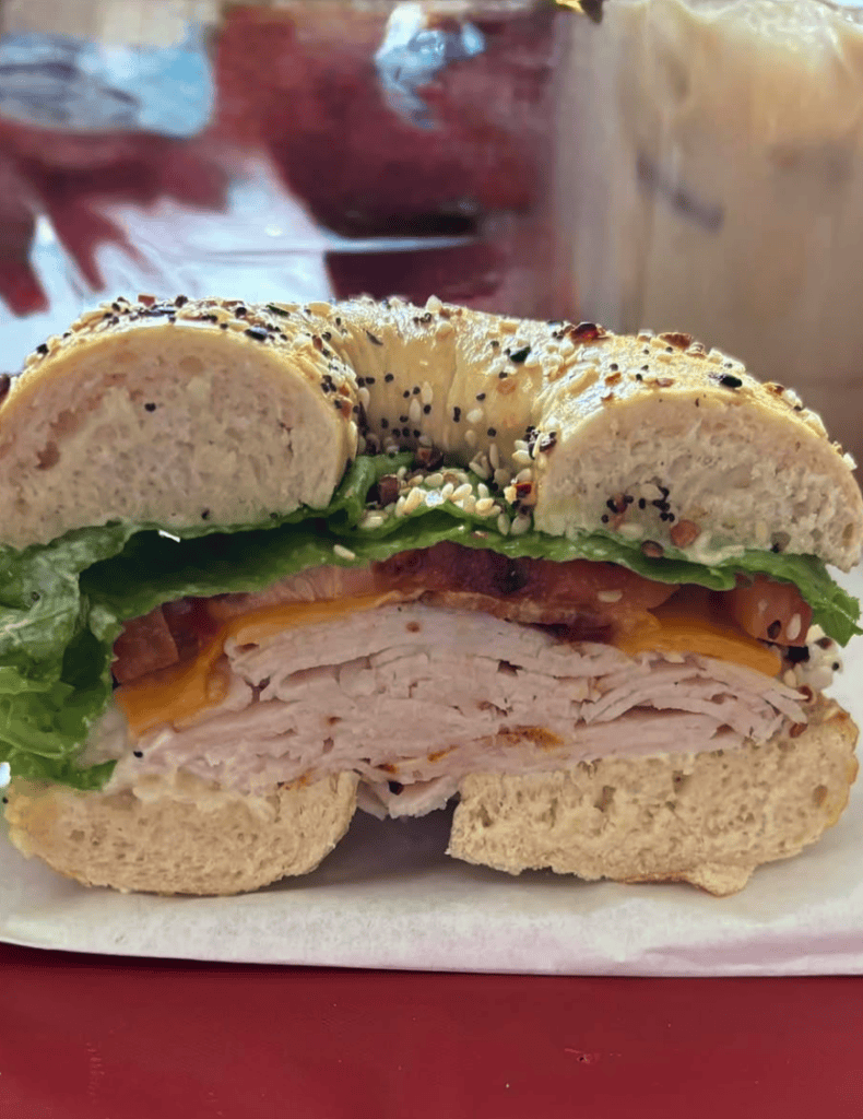 A close-up of a fresh bagel sandwich filled with sliced ham and lettuce, served on a white plate with beverages in the background.