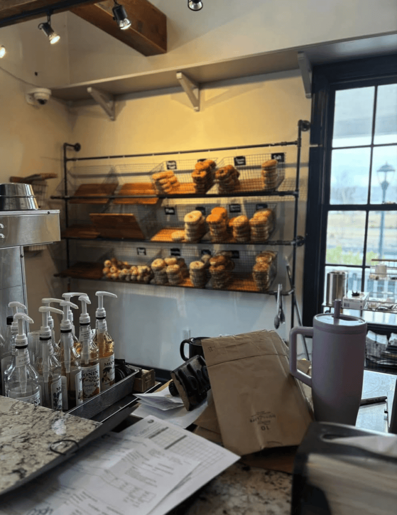 A modern bakery interior features a wall-mounted display of baked goods and pastries with a service counter in the foreground and large windows showing an urban street view.
