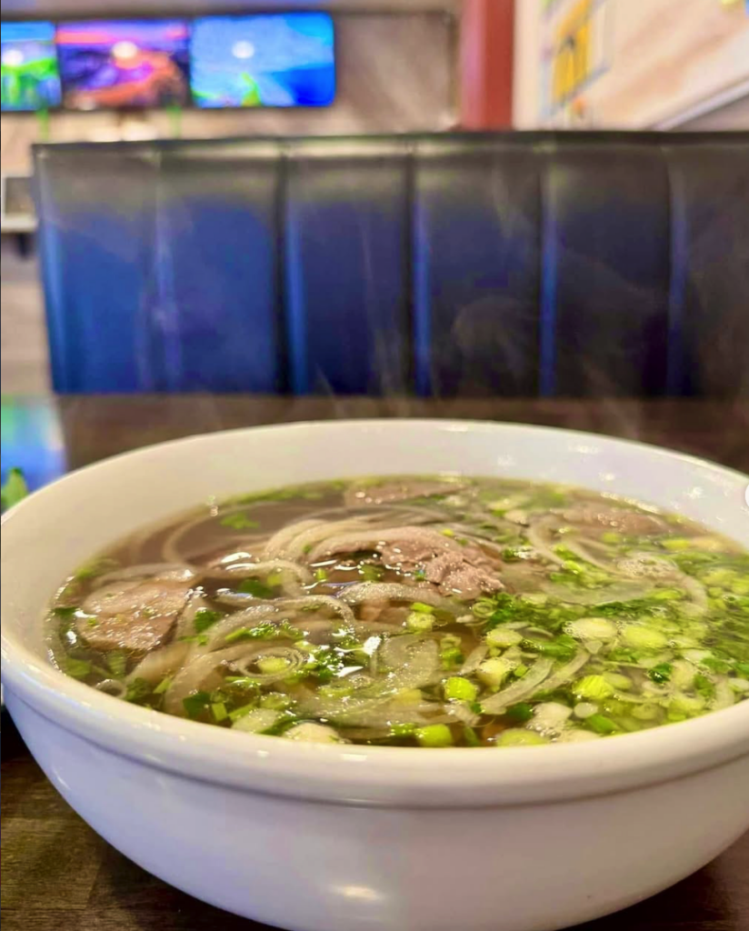 A close-up of a steaming bowl of noodle soup with meat and green vegetables in a white bowl, served in a casual restaurant setting.