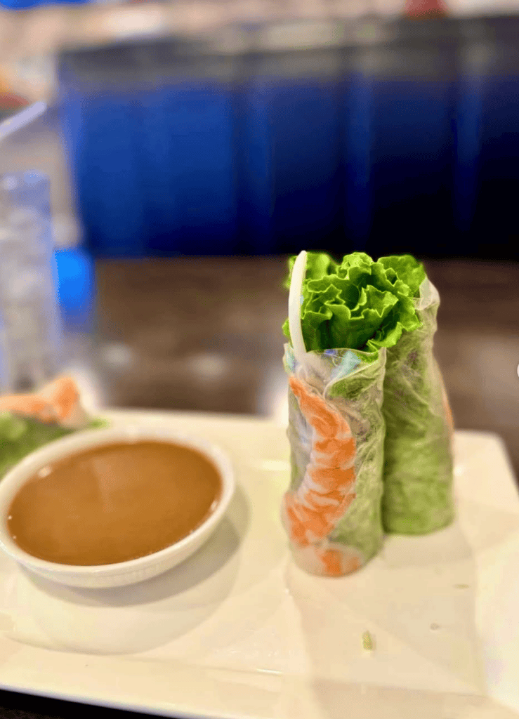 A freshly prepared spring roll filled with vegetables is presented on a white plate alongside a small bowl of peanut dipping sauce in a restaurant setting.