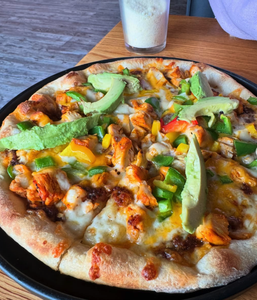 An overhead view of a freshly prepared pizza topped with vegetables and melted cheese on a wooden table with a beverage nearby.