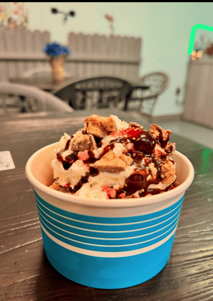 A close-up shot of a creamy ice cream sundae topped with caramel and chocolate bits served in a blue striped paper cup at an indoor venue.