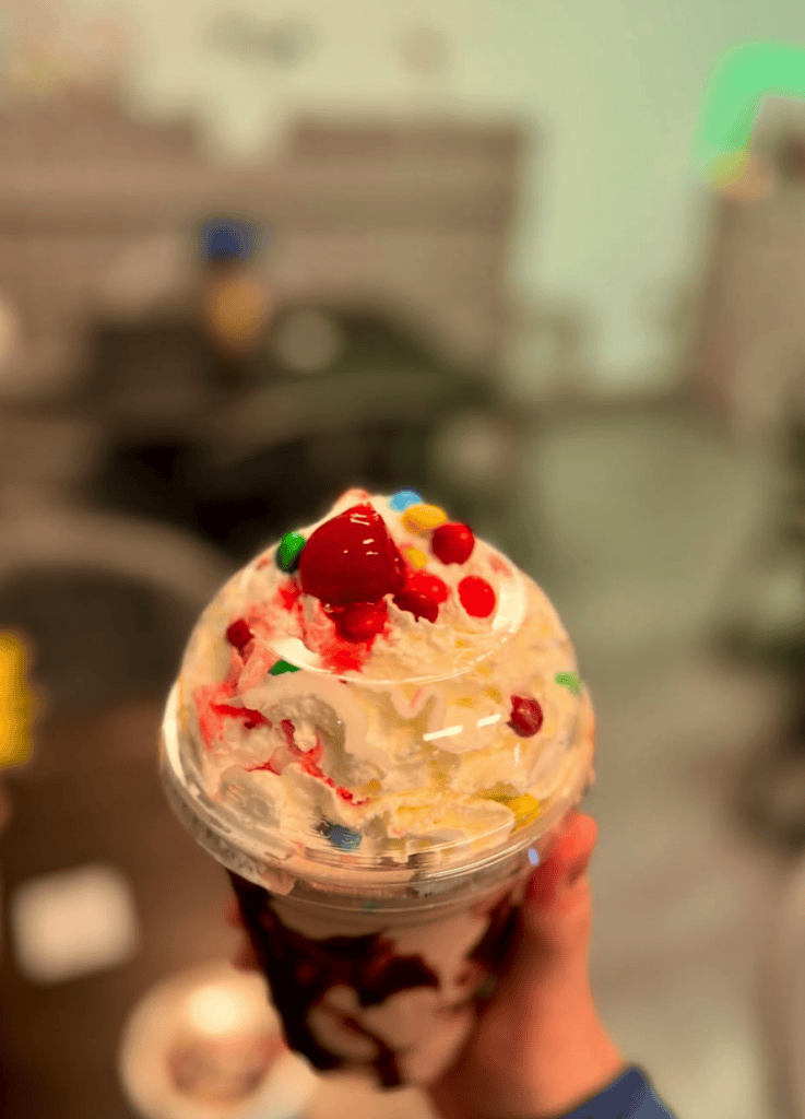 A close-up shot of a colorful ice cream sundae topped with whipped cream and red sprinkles being held up in someone's hand.