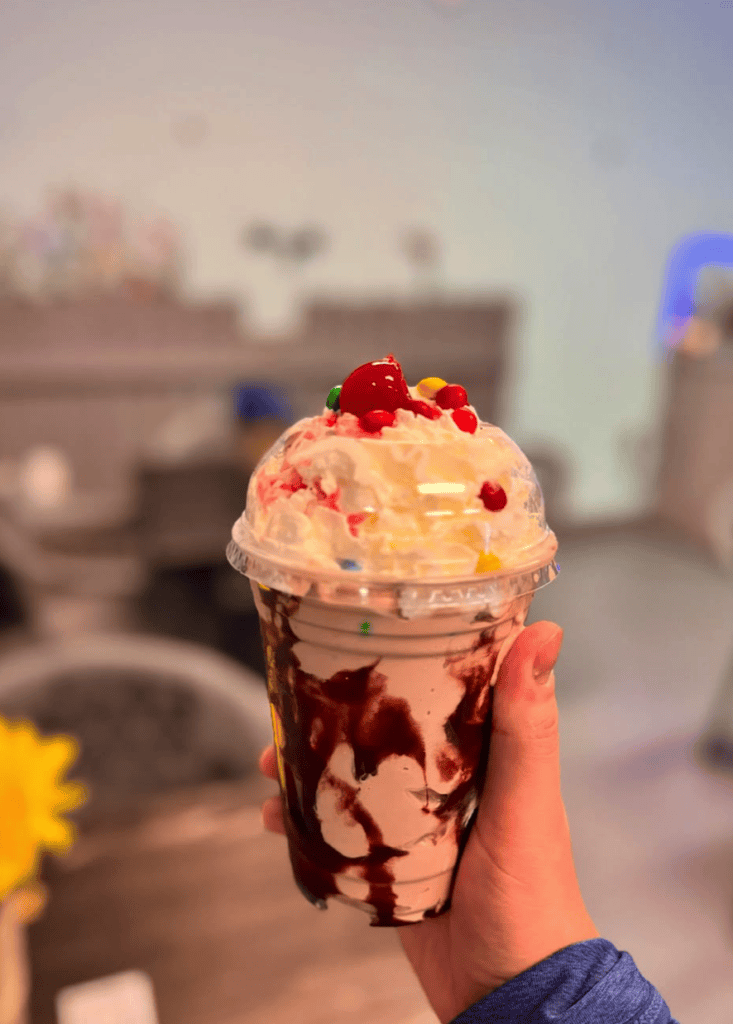 A hand holds an elaborate ice cream sundae with whipped cream, chocolate sauce, and red cherries against a blurred urban outdoor background.