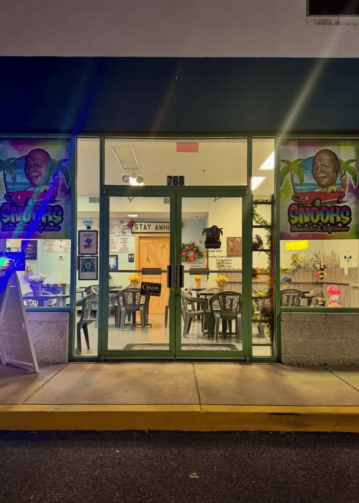 A brightly lit storefront venue with neon signage and glass doors opens onto a dark sidewalk, showcasing an interior space with seating and merchandise displays.