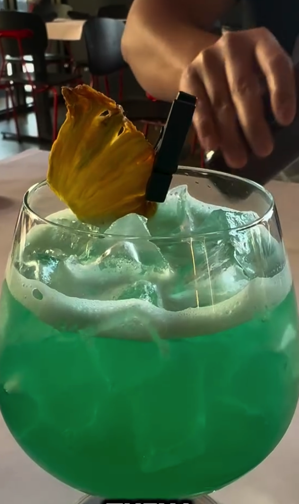 A person pours a vibrant green cocktail into a glass filled with ice, with text overlay reading 'THEN I GOT THIS'.