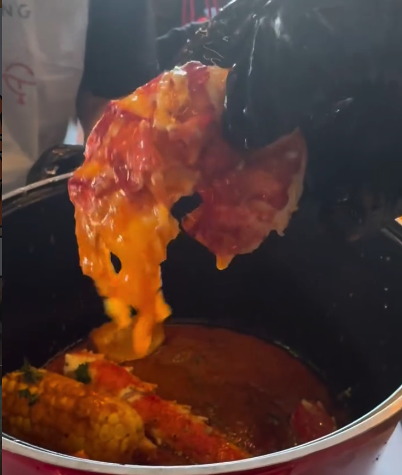 A close-up shot of meat being cooked in a pot with tongs lifting a piece above steaming liquid, capturing the cooking process with rich amber and orange tones.