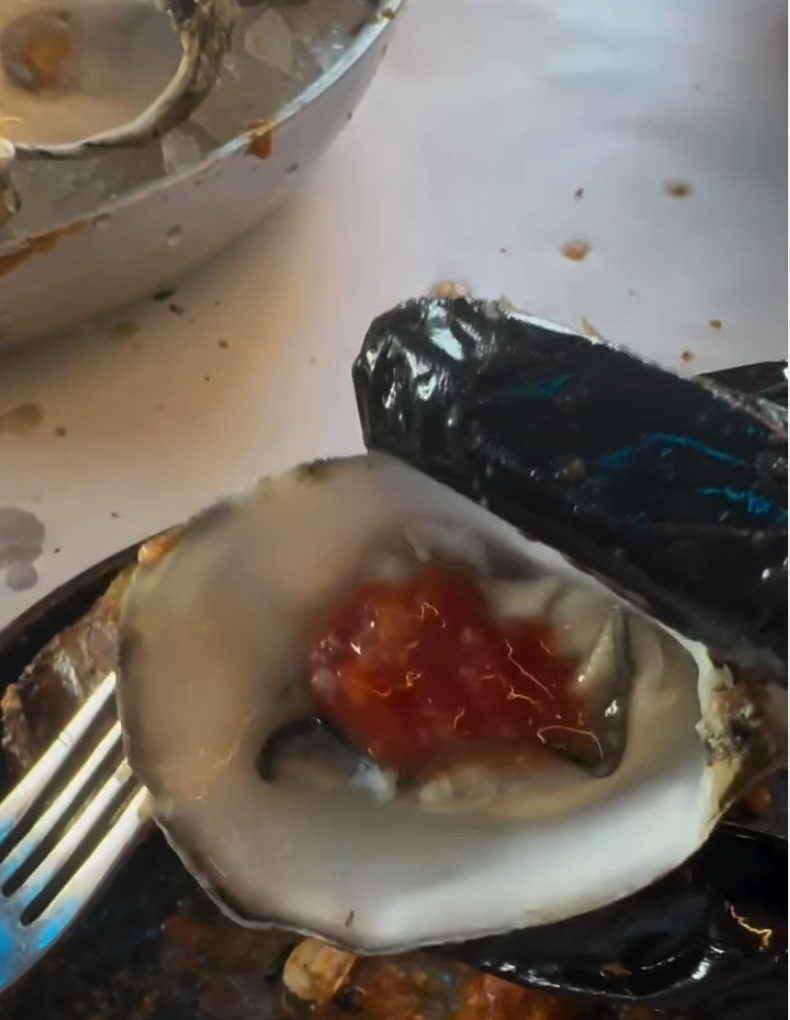 A fresh oyster on the half shell topped with red sauce, presented on a white plate with dining utensils visible in the frame.