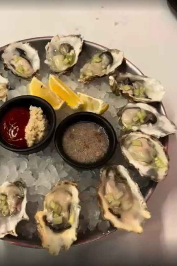 A beautifully arranged platter of fresh oysters on ice with lemon wedges and two dipping sauces presented overhead.