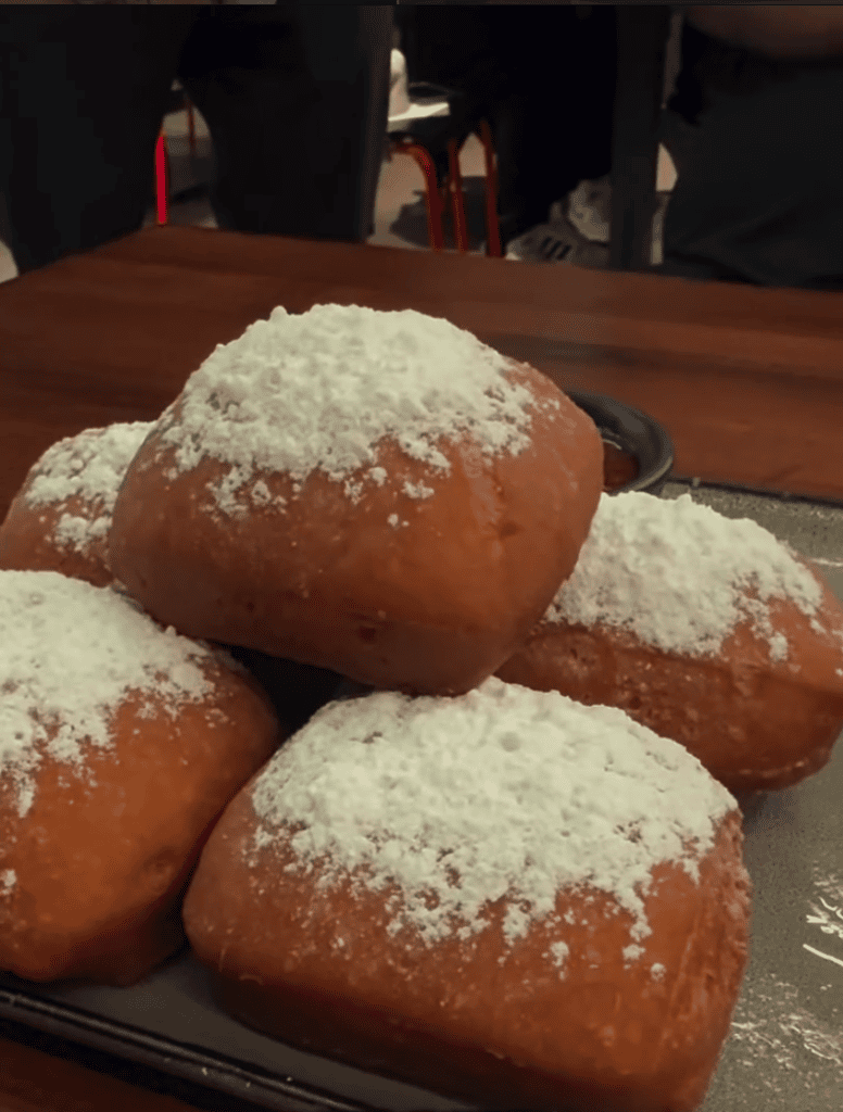 Four golden-fried donuts dusted with powdered sugar arranged on a black plate on a wooden table at what appears to be an indoor venue or restaurant.