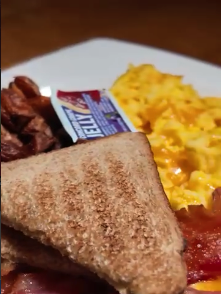 A close-up photograph of a breakfast plate featuring scrambled eggs, crispy bacon, wheat toast, and a butter packet on a white plate.