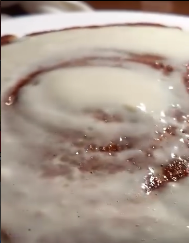 A close-up overhead view of a freshly glazed cinnamon roll with white frosting drizzle on a ceramic plate.