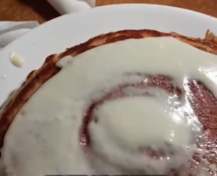 A close-up overhead view of a freshly frosted cinnamon roll on a white plate with a wooden surface visible in the background.