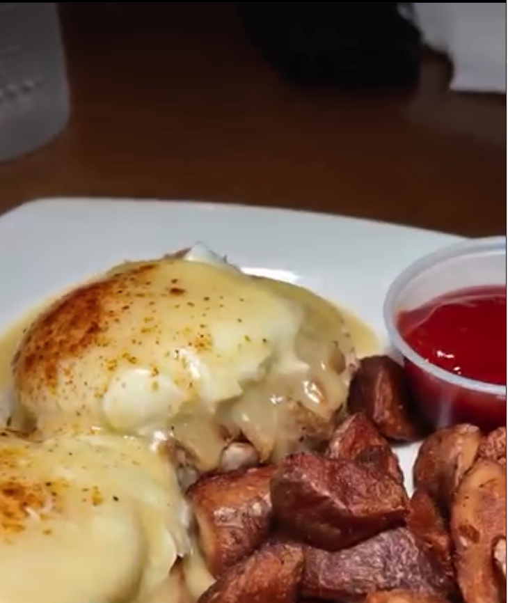 A close-up overhead shot of a loaded baked potato with melted cheese, crispy bacon, and sour cream, served on a white plate with a side of red sauce.