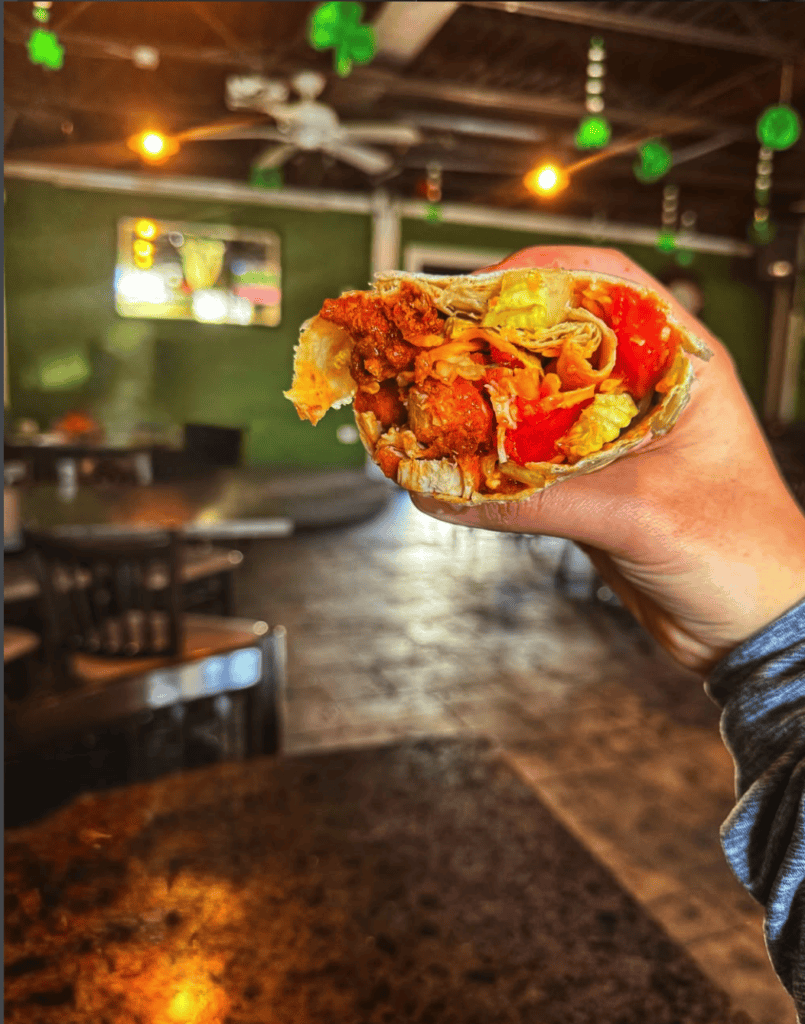 A hand holds up a colorful waffle sandwich filled with various toppings in what appears to be a casual restaurant or food establishment.