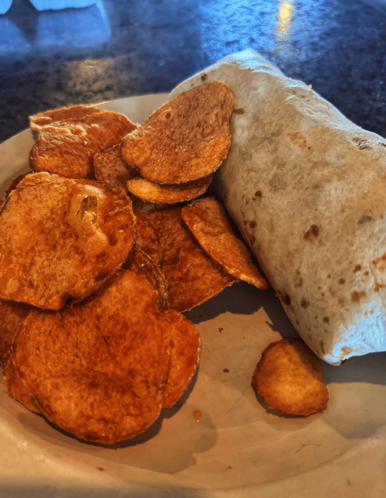 A close-up overhead shot of a plated meal featuring crispy sweet potato chips, a wrapped sandwich or burrito, and other fried accompaniments on a light-colored plate.