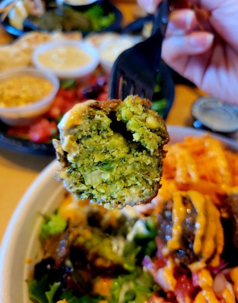 A close-up shot of a vibrant pistachio-crusted falafel being held above a colorful Middle Eastern or Mediterranean food platter with various dishes in the background.