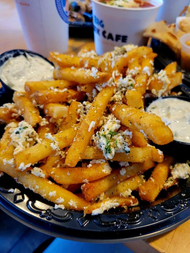 A close-up overhead shot of crispy golden french fries topped with cheese, seasonings, and garnishes, served on a dark plate with dipping sauce on the side.