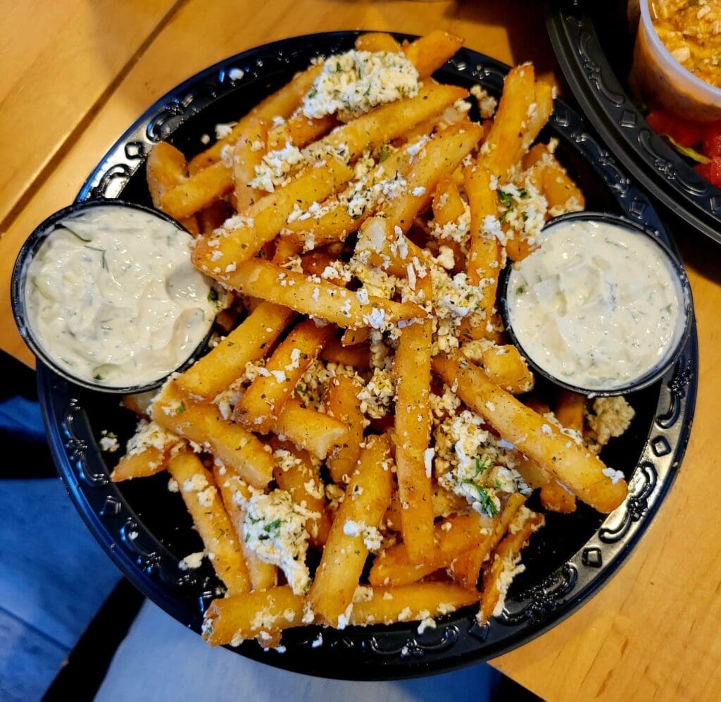 A black plate overflowing with golden crispy fries topped with melted cheese, herbs, and seasonings, served with two white dipping sauce cups on the sides.