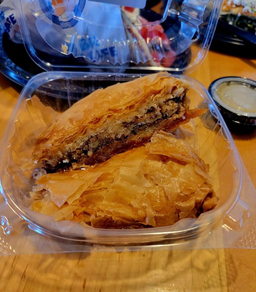 A golden fried sandwich and pastry served in a disposable container with a side of dipping sauce and beverage visible on a wooden surface.
