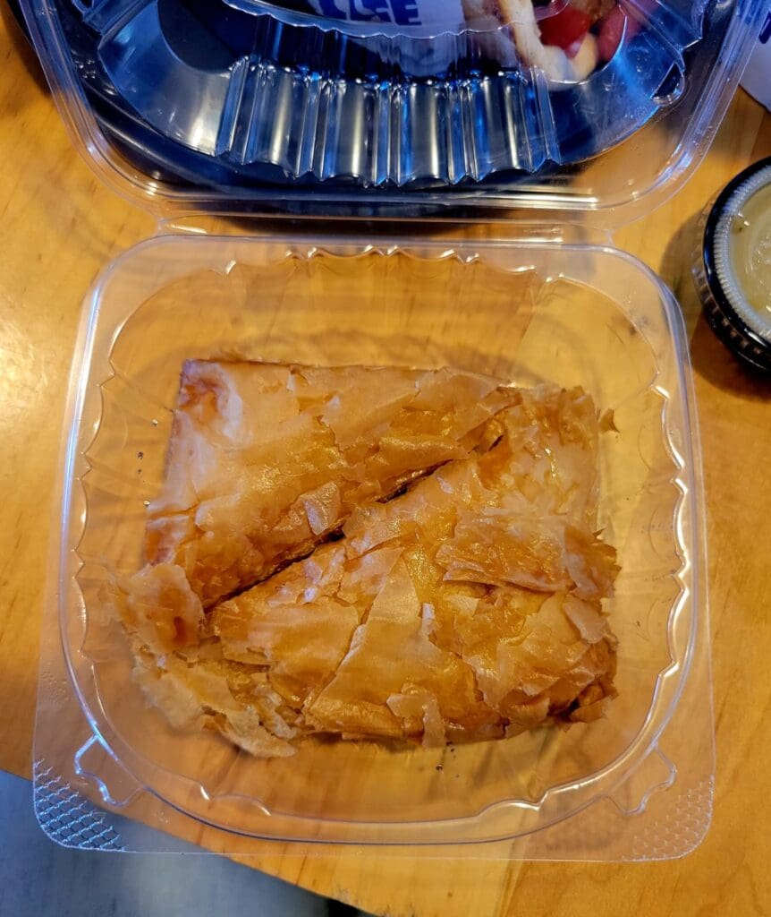 A golden-brown baked pastry item sits in a clear plastic takeout container on a wooden surface.