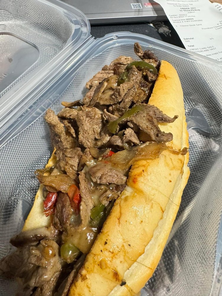 A close-up overhead view of a Philly cheesesteak sandwich topped with sautéed onions and peppers in a clear plastic container.