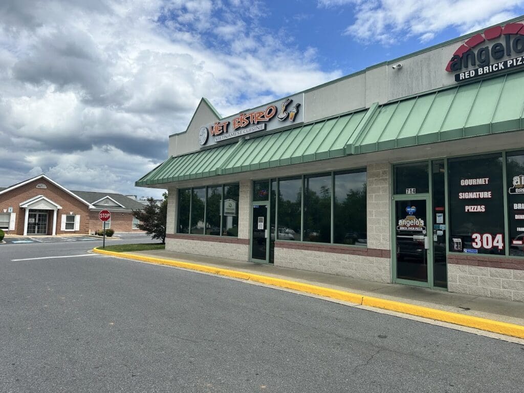 A small commercial storefront with a green metal roof and awning sits along a quiet parking lot on a partly cloudy day.
