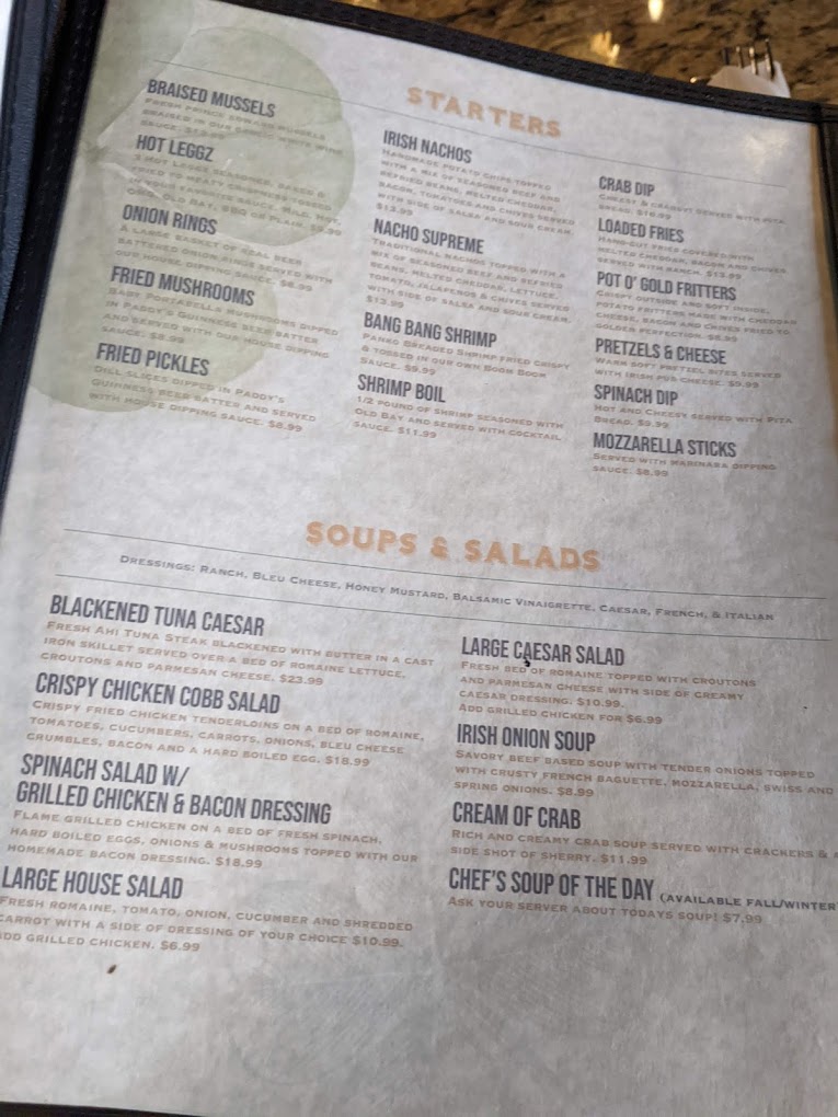 A close-up overhead view of a restaurant menu displayed in a frame, showing organized sections for soups, salads, and other dishes with cream and beige tones.