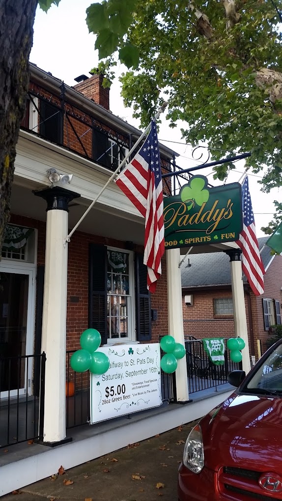 A charming storefront with an American flag hangs above the entrance, featuring green signage and white trim, located on a quiet street with parked cars and tree shade.