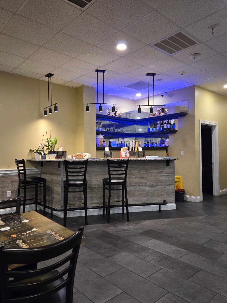 A modern restaurant or bar interior features a sleek black counter with blue accent lighting on shelving behind it, pendant lights overhead, and dining chairs in the foreground.